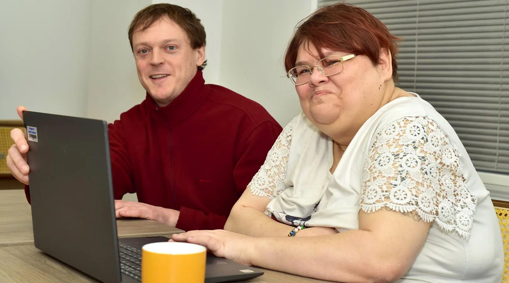 Two people seated together at a table, smiling and using a laptop.